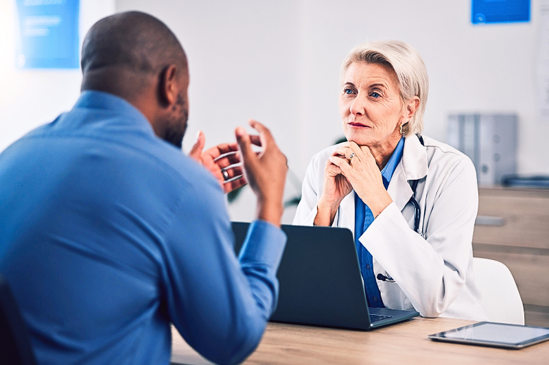 A mature female doctor attentively listens to a male patient during a consultation in a modern medical office. She rests her chin on her hands and maintains thoughtful eye contact, while the patient gestures as he speaks. A laptop and tablet sit on the desk between them.